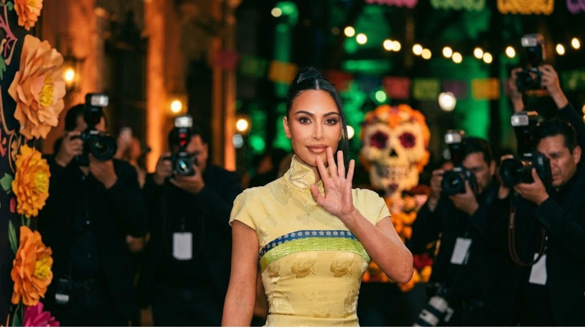 Kim Kardashian saluda en un evento nocturno en México, rodeada de fotógrafos y decoración cultural desenfocada.